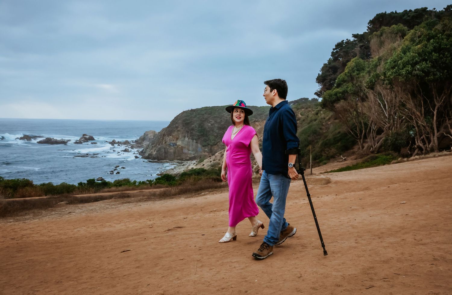 Couple walking on coastal trail, woman in pink dress and man with cane, scenic ocean cliffs in background.