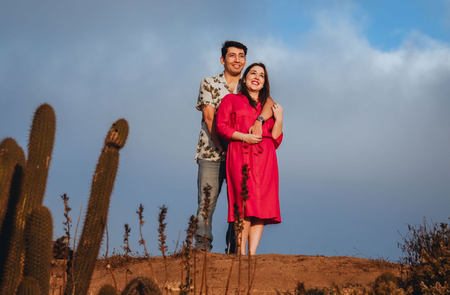 Couple embracing on red desert hill with cacti under blue cloudy sky in romantic outdoor photo session.