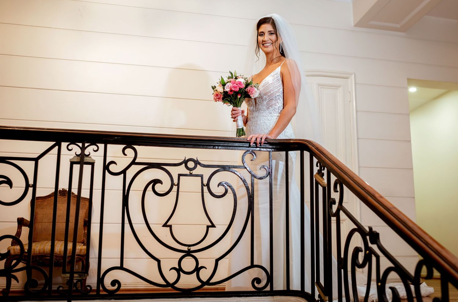 Bride in white lace gown holding pink bouquet poses gracefully on ornate black iron staircase railing.