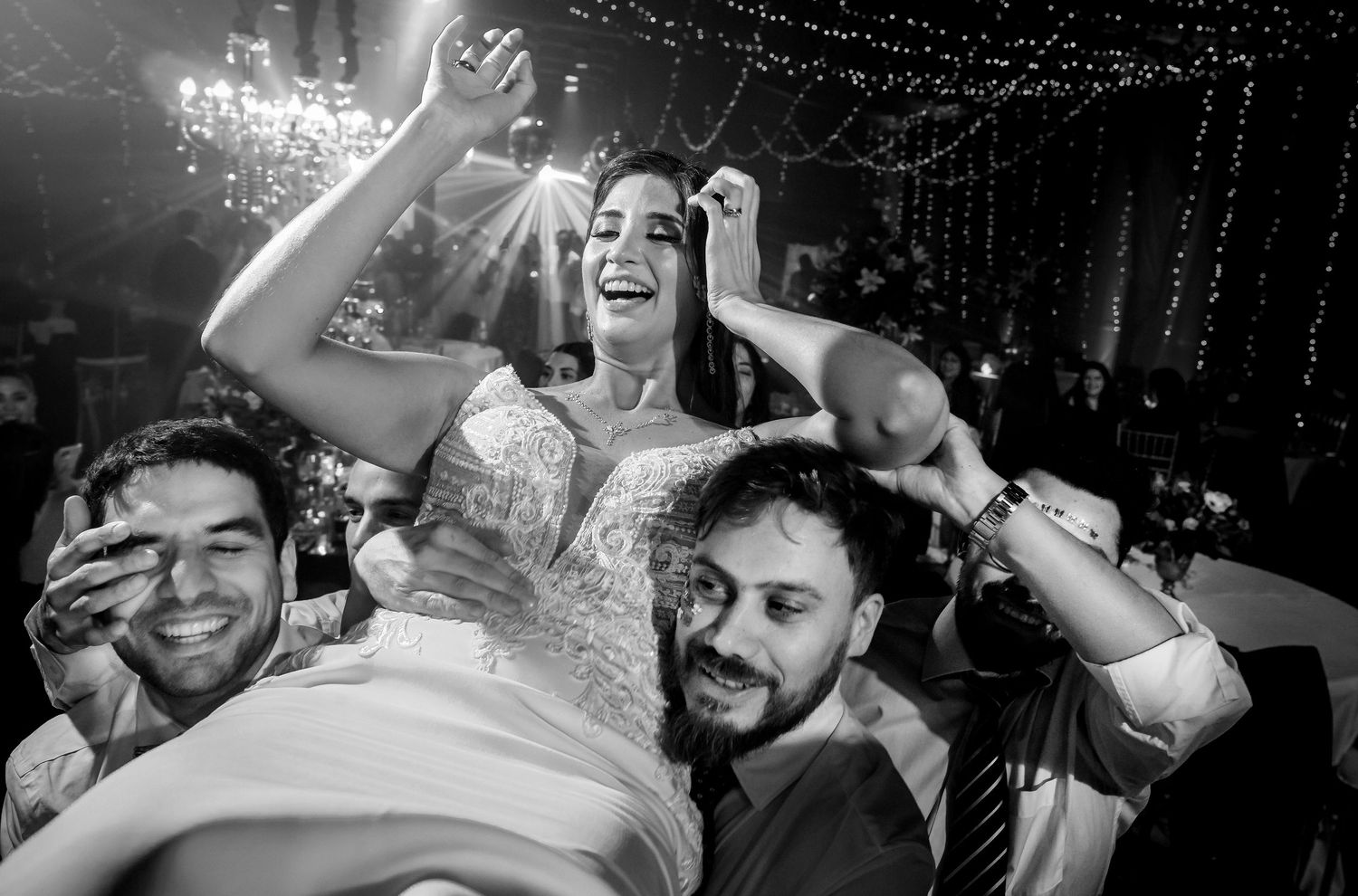 Bride laughing joyfully while being lifted by guests during a lively black and white wedding reception celebration.