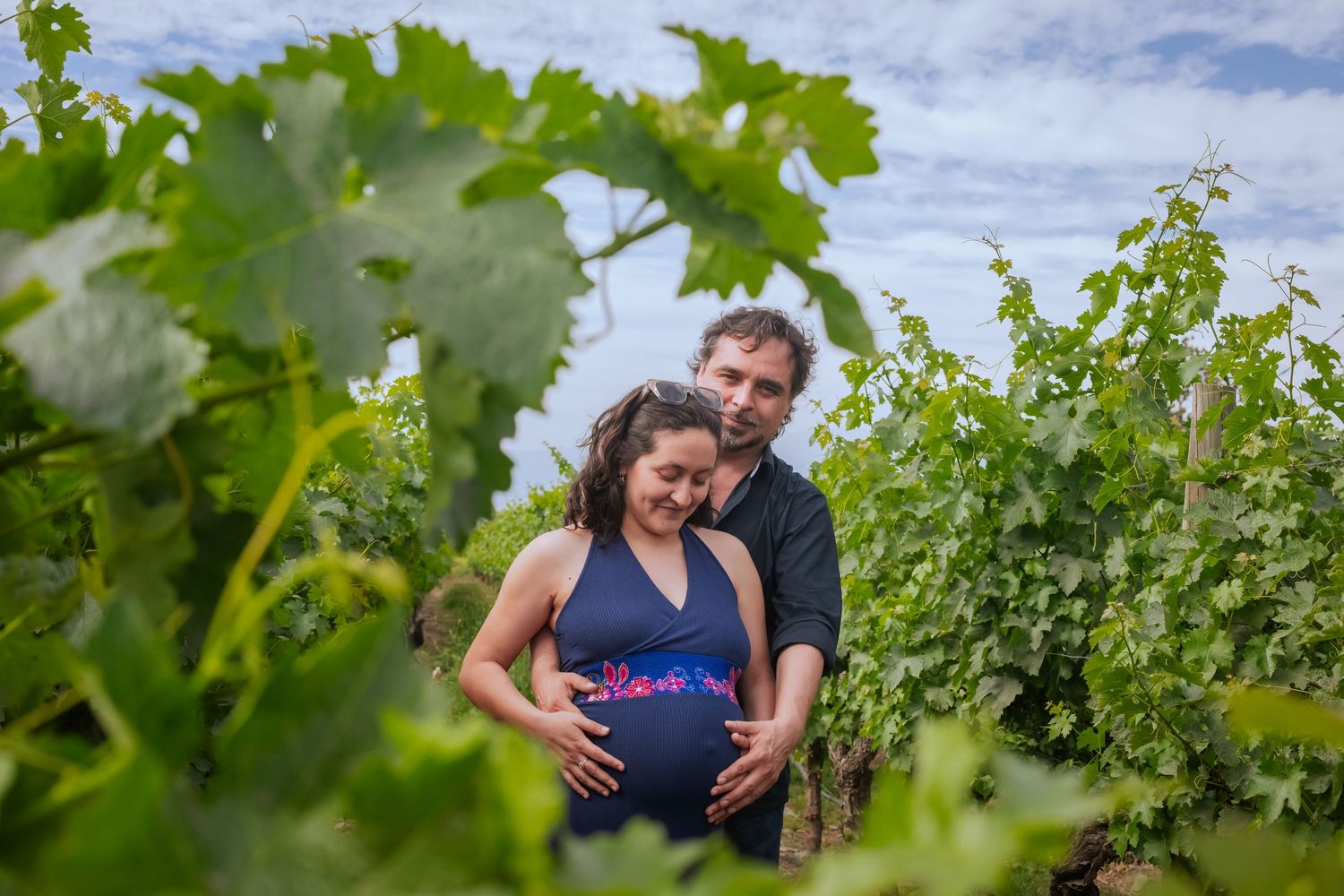 Couple embracing among green grapevines in vineyard, man tenderly holding pregnant partners belly.