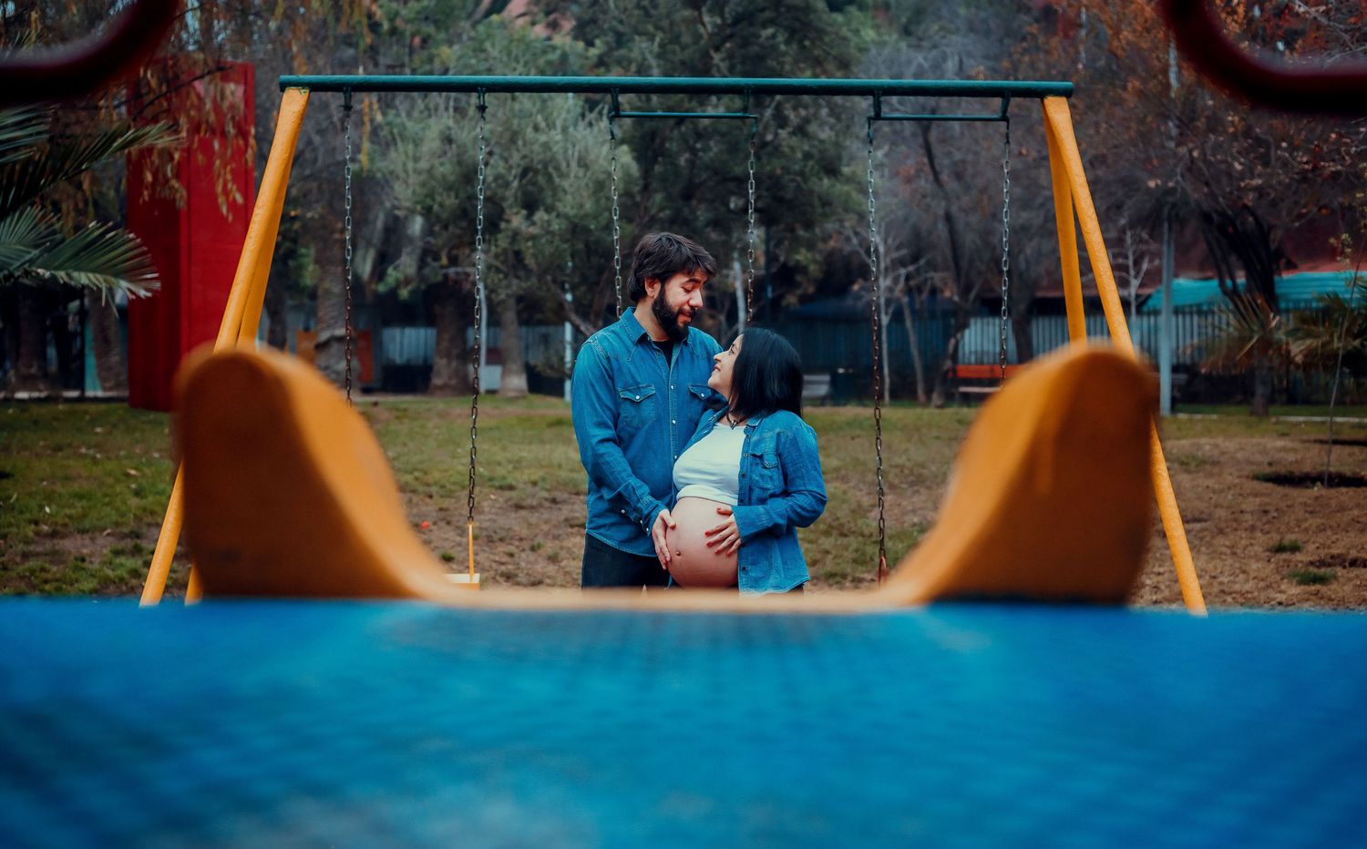 Couple embracing at playground, man holding pregnant woman's baby bump near yellow swing set outdoors.