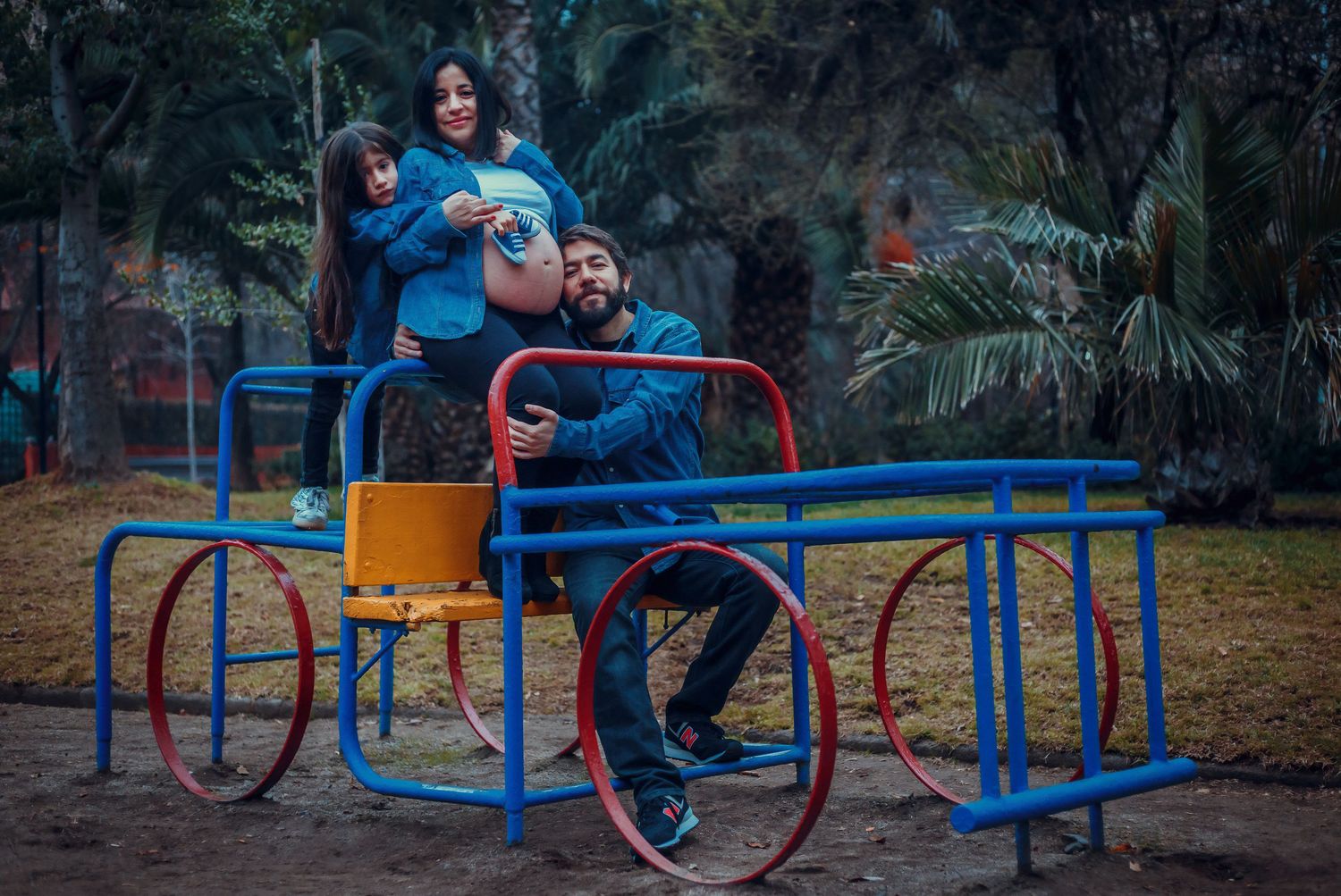 Family playing on a merry-go-round at a park, pregnant mother and child sitting on man.