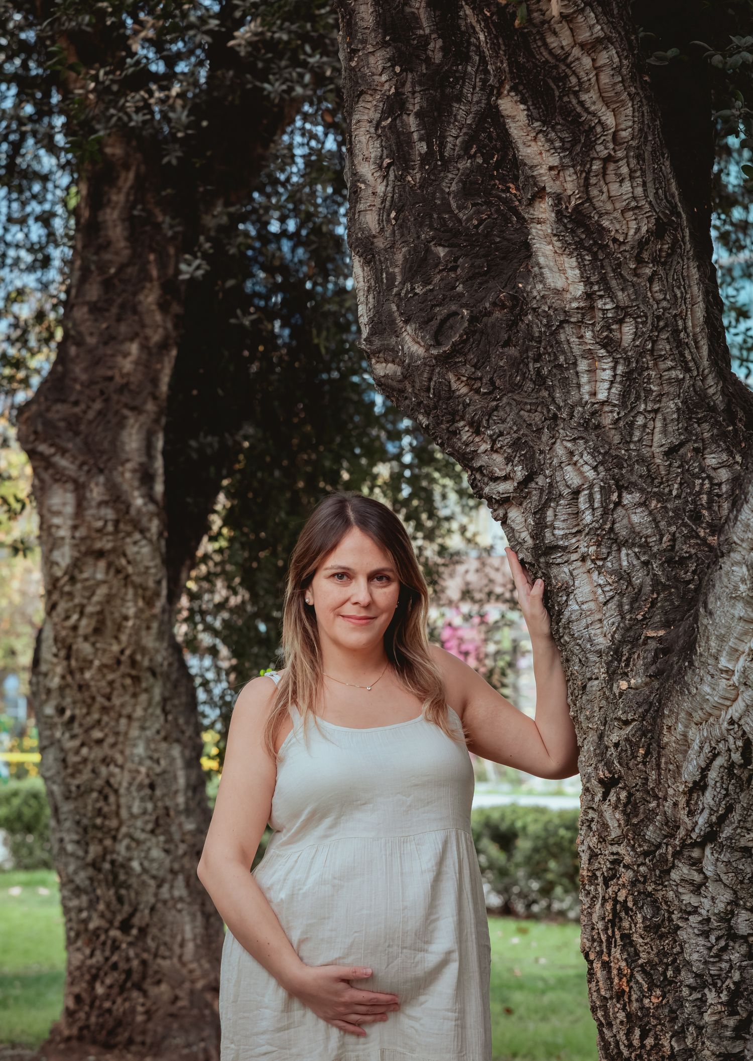 Smiling pregnant woman in white dress posing between two large trees in a sunny park.