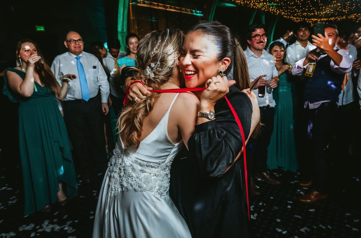 Bride and guest share a joyful hug on the wedding dance floor surrounded by celebrating guests.