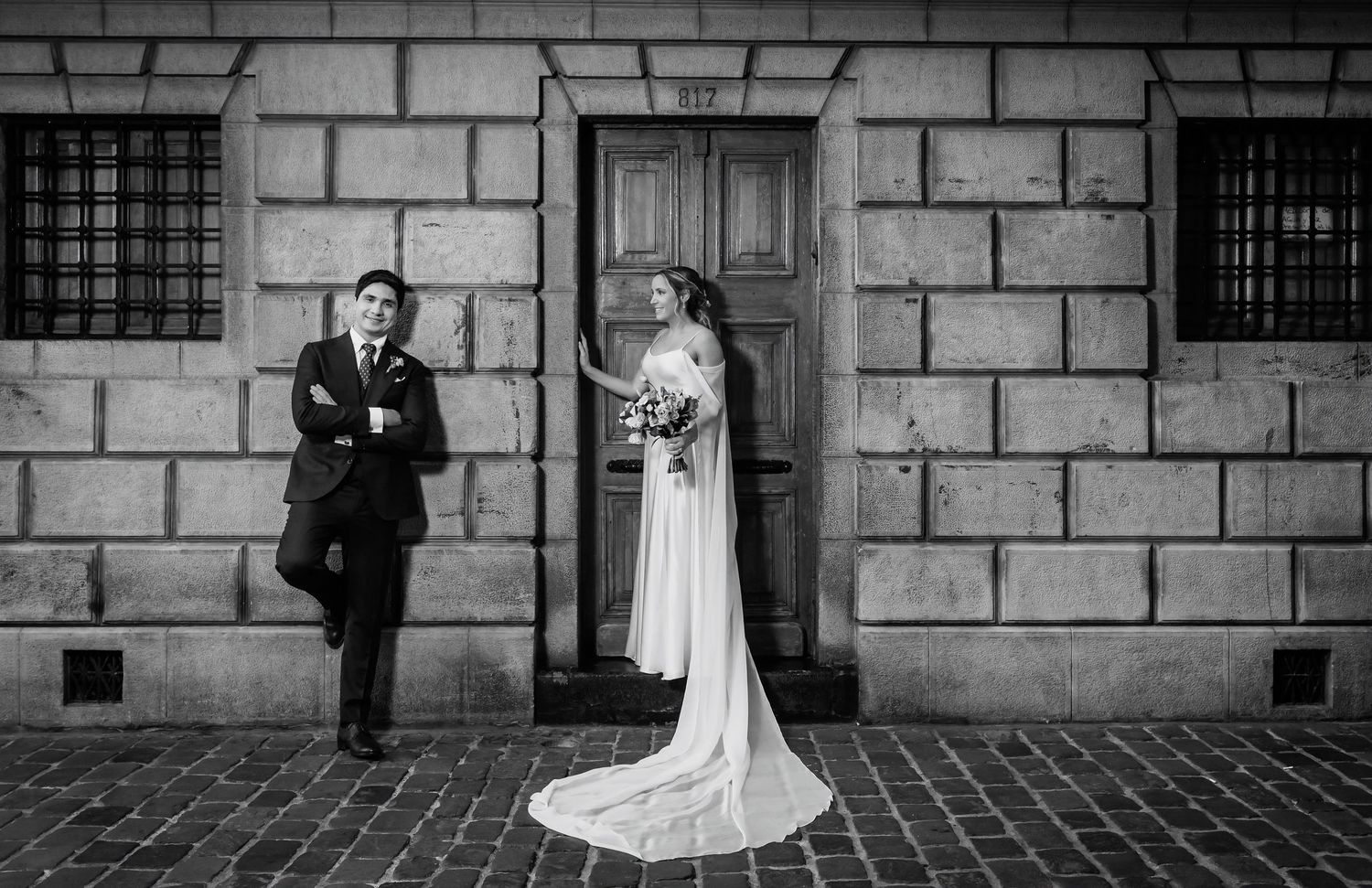Black and white photo of bride and groom posing separately outside stone building with wooden door.