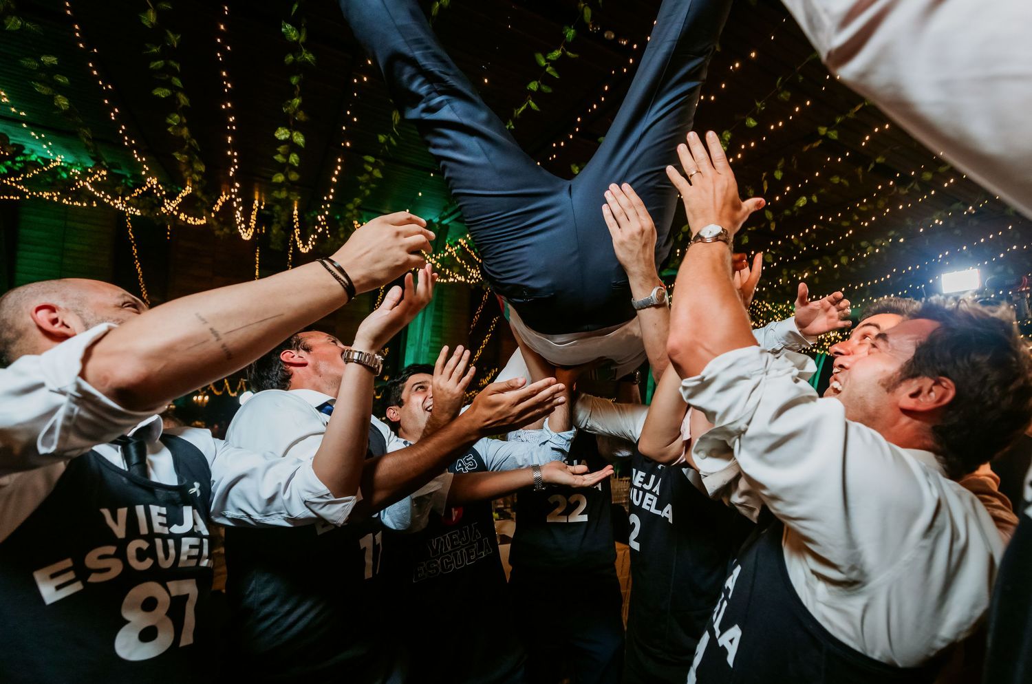 Groomsmen in matching jerseys lifting and crowd surfing guest at energetic wedding reception.
