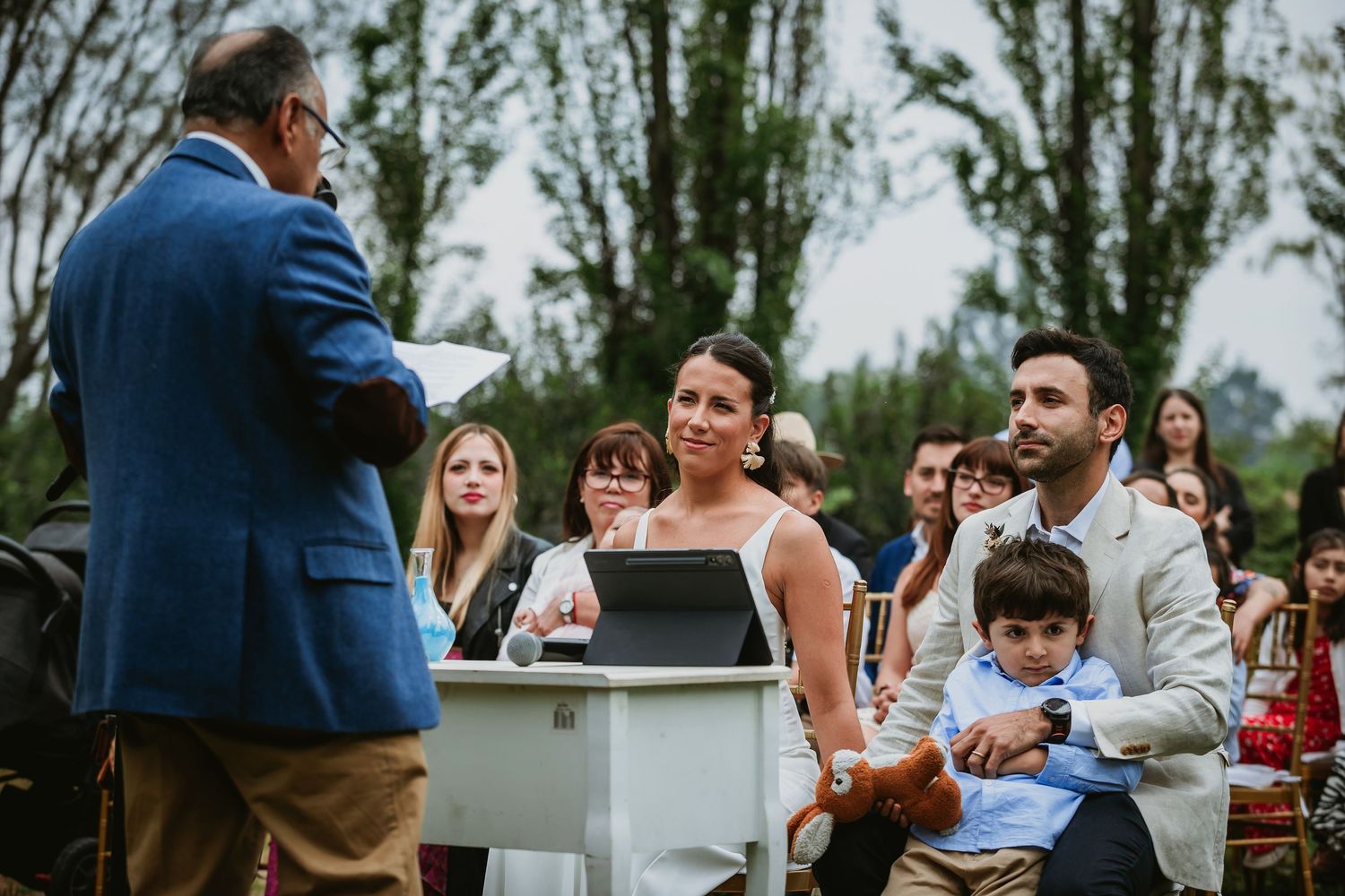 Bride and groom at outdoor wedding ceremony with officiant at podium and guests seated in background.