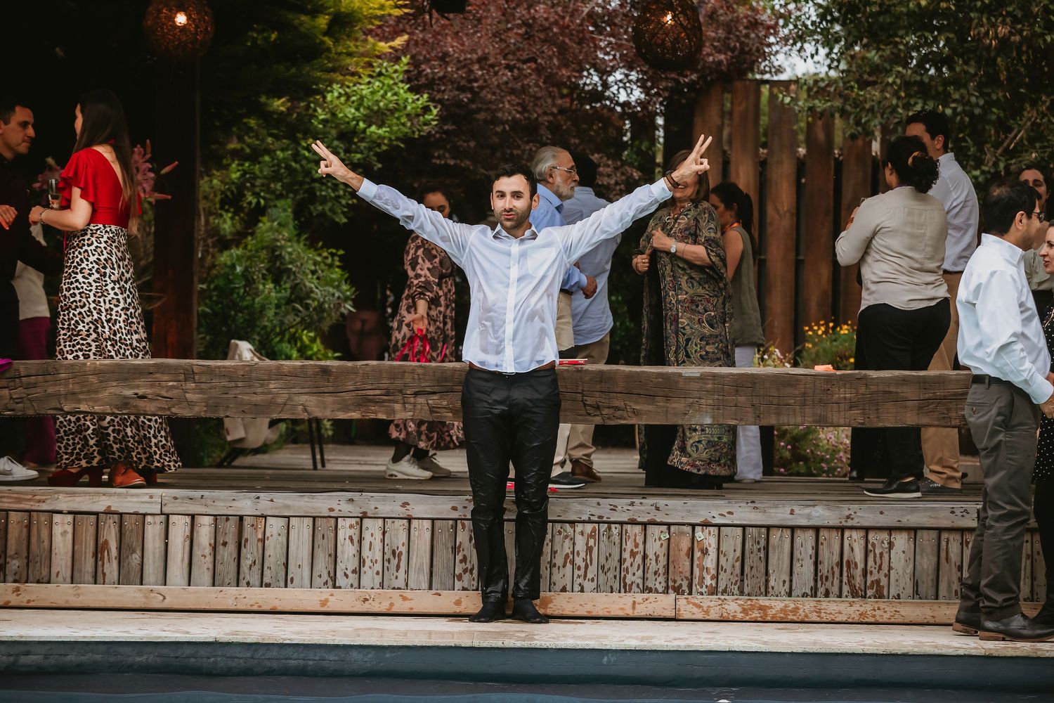 Bearded man in white shirt raises arms wide at an outdoor party celebration with guests mingling in background.