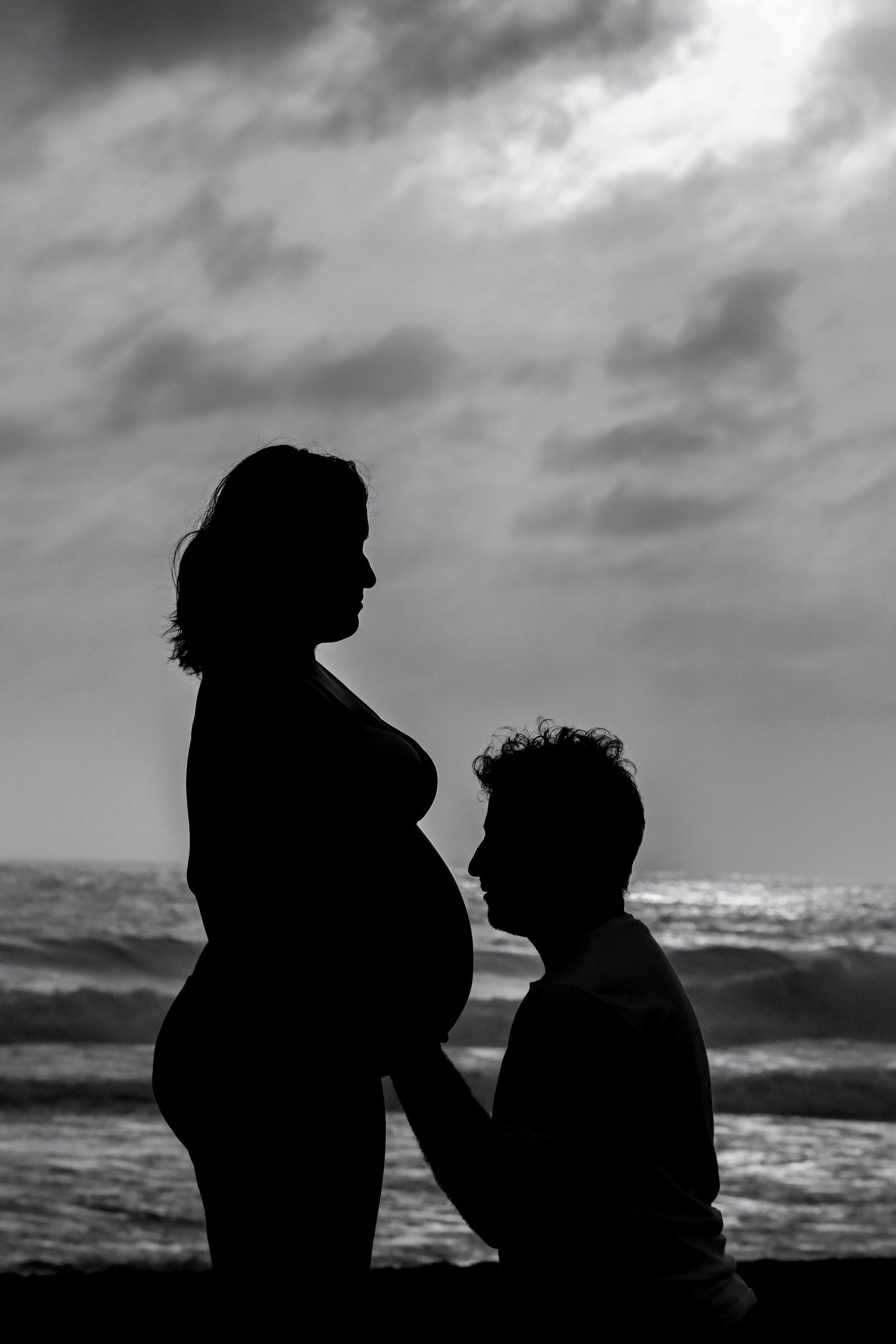 Black and white silhouette of pregnant woman and partner kneeling before her belly at ocean beach.