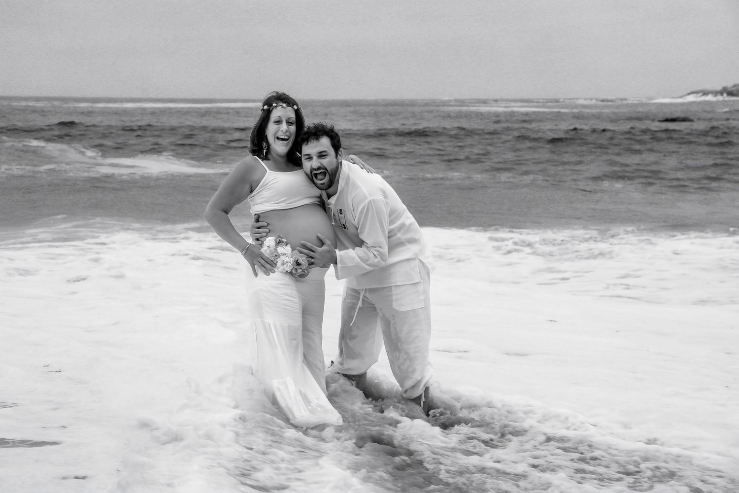 Black and white maternity photo of couple laughing in ocean waves, pregnant woman in flowing white dress.