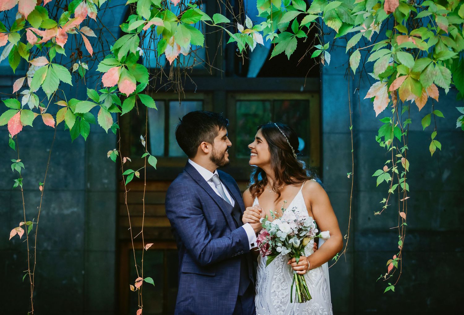 A couple shares a romantic moment under colorful autumn vines, bride in lace gown holding bouquet, groom in navy suit.