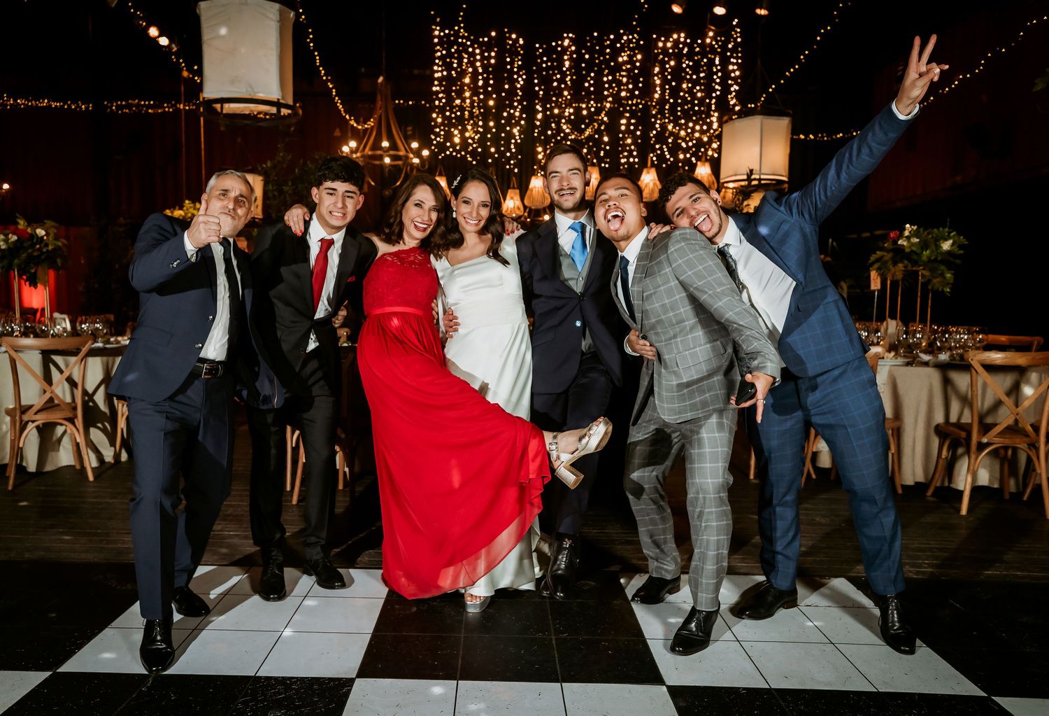Wedding couple poses with groomsmen on checkered dance floor at elegant reception with string lights.