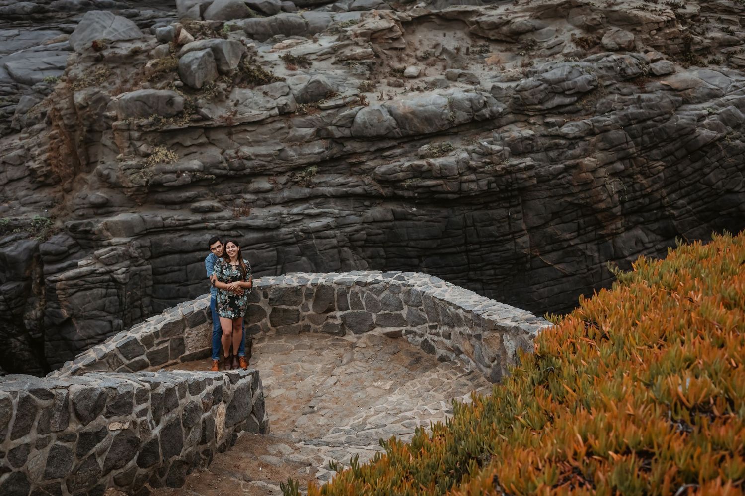 Couple standing together on stone bridge surrounded by rocky cliffs and autumn foliage.