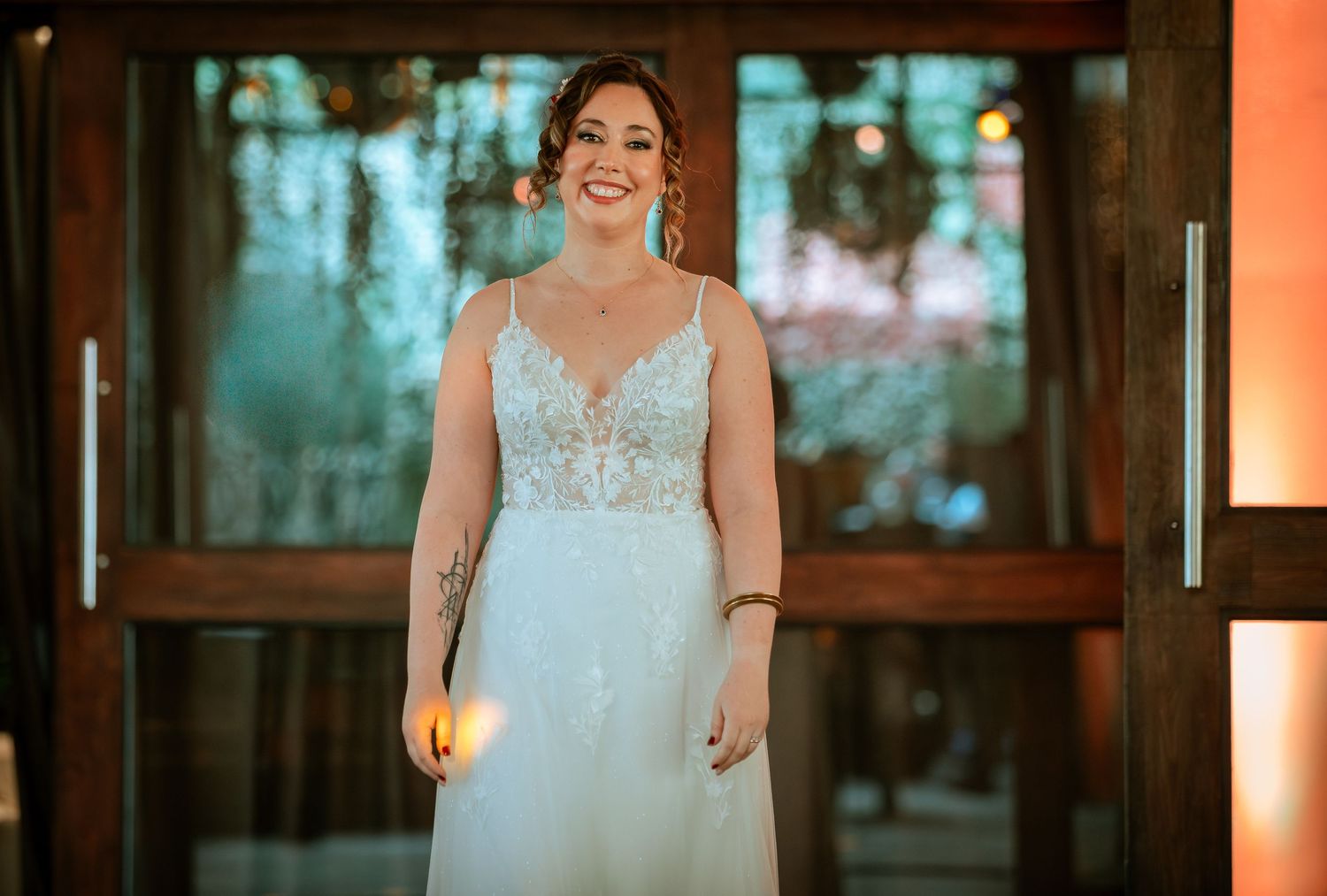 Smiling bride in elegant lace wedding gown stands in warmly lit wedding venue doorway.