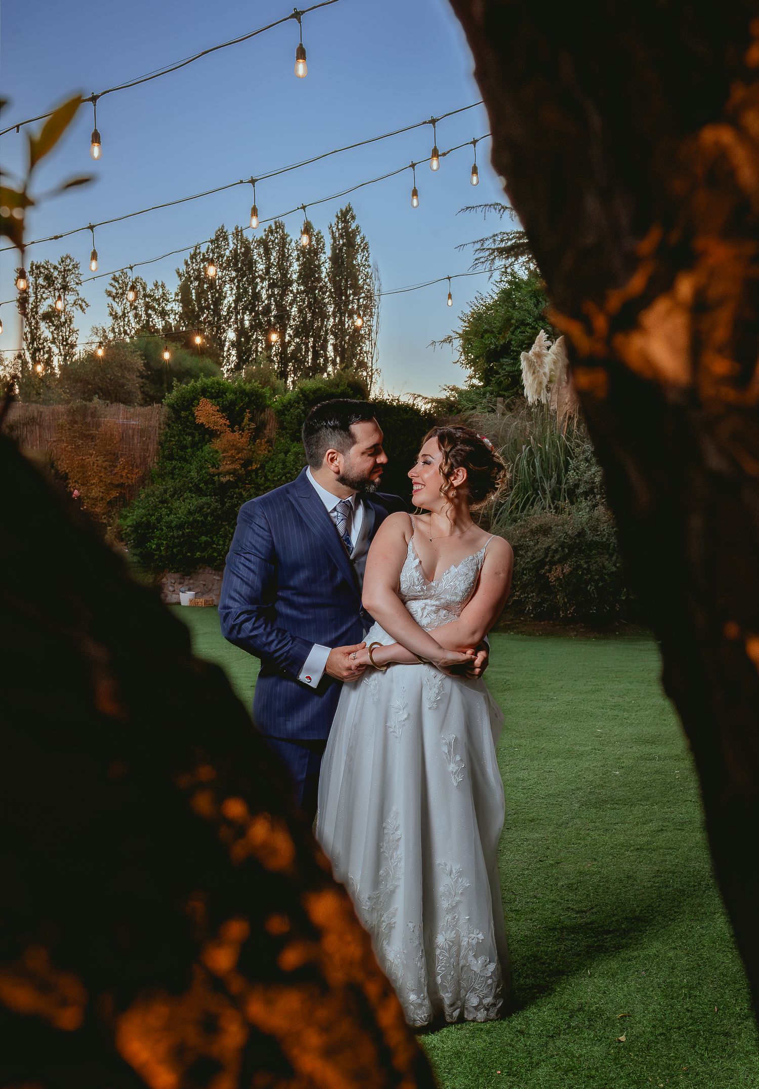 Bride and groom share a romantic moment on green lawn under string lights at dusk wedding.
