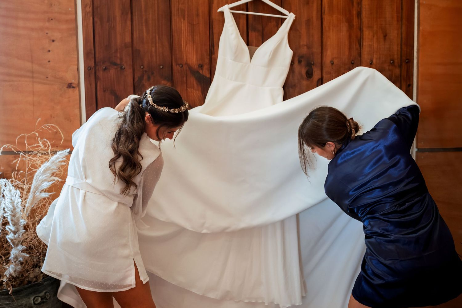 Two women in robes carefully handling a white wedding gown hanging on a wooden wall.