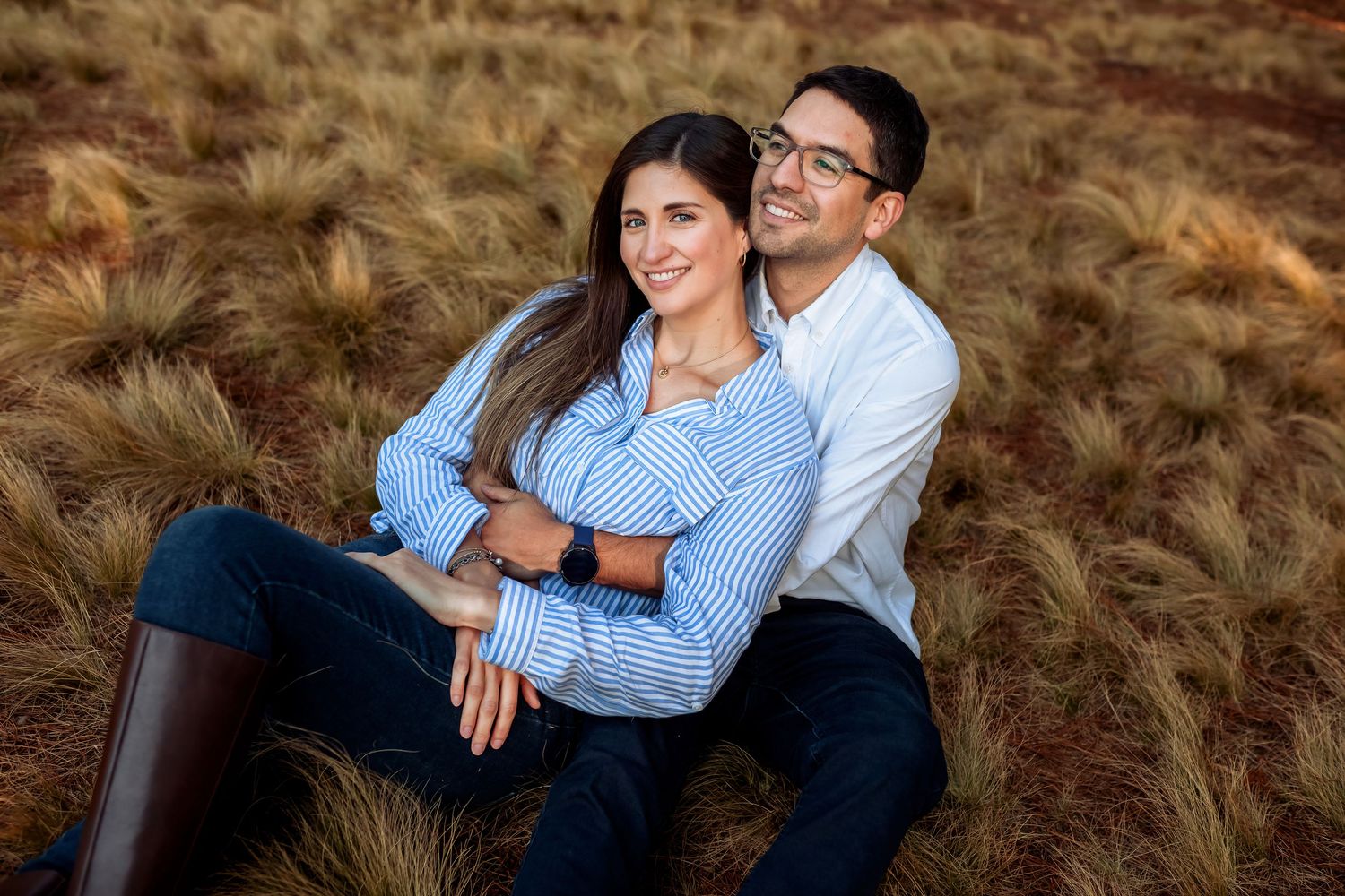 A couple sitting together in a field of golden dry grass, smiling warmly at the camera.