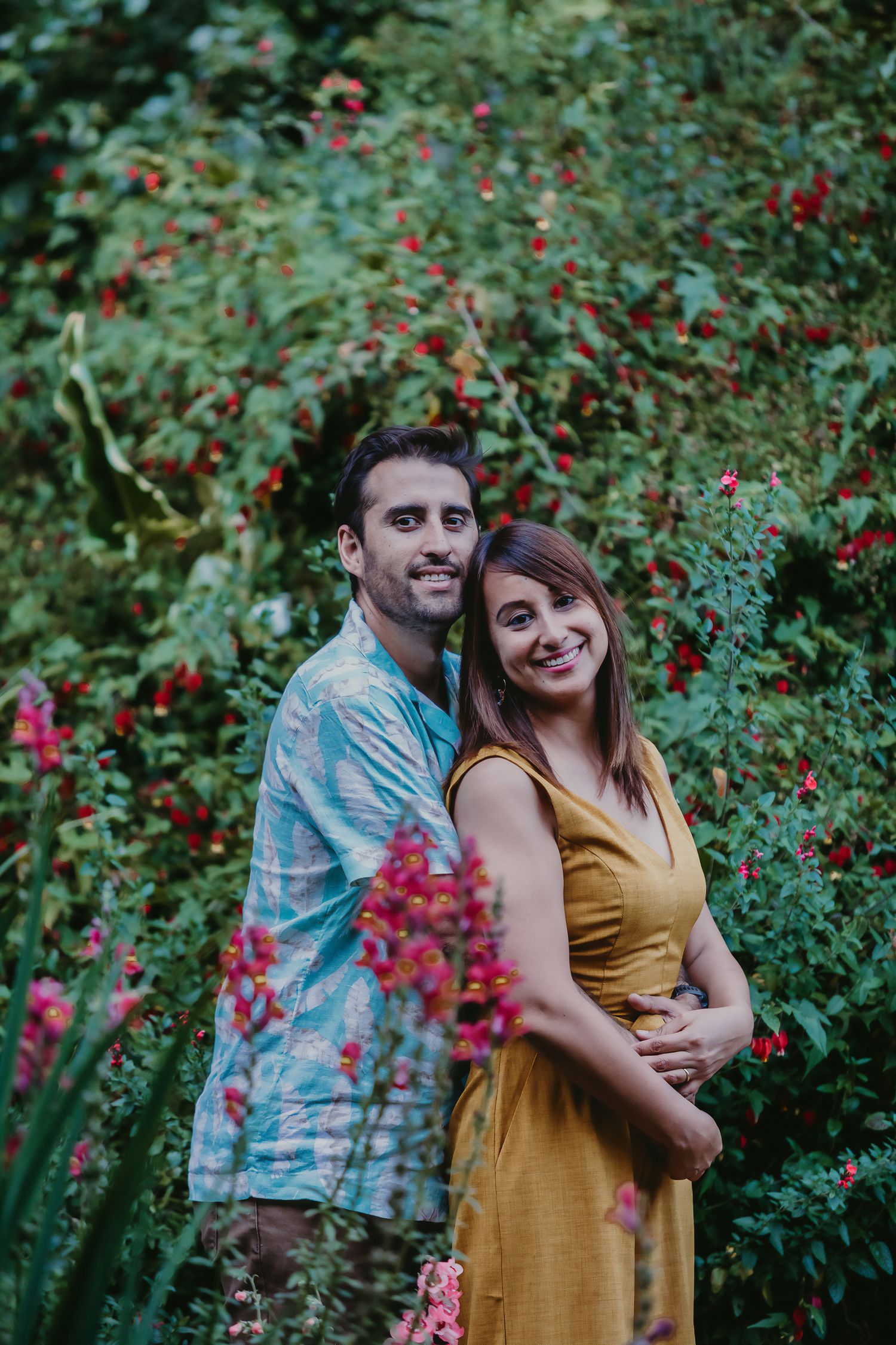 Smiling couple posing together in front of lush green foliage and red flowers in a garden.