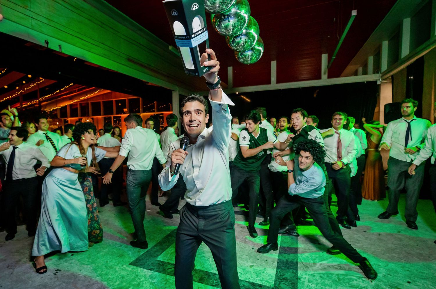 Excited man holds up a shoe at a festive wedding reception with green balloons and dancing guests.