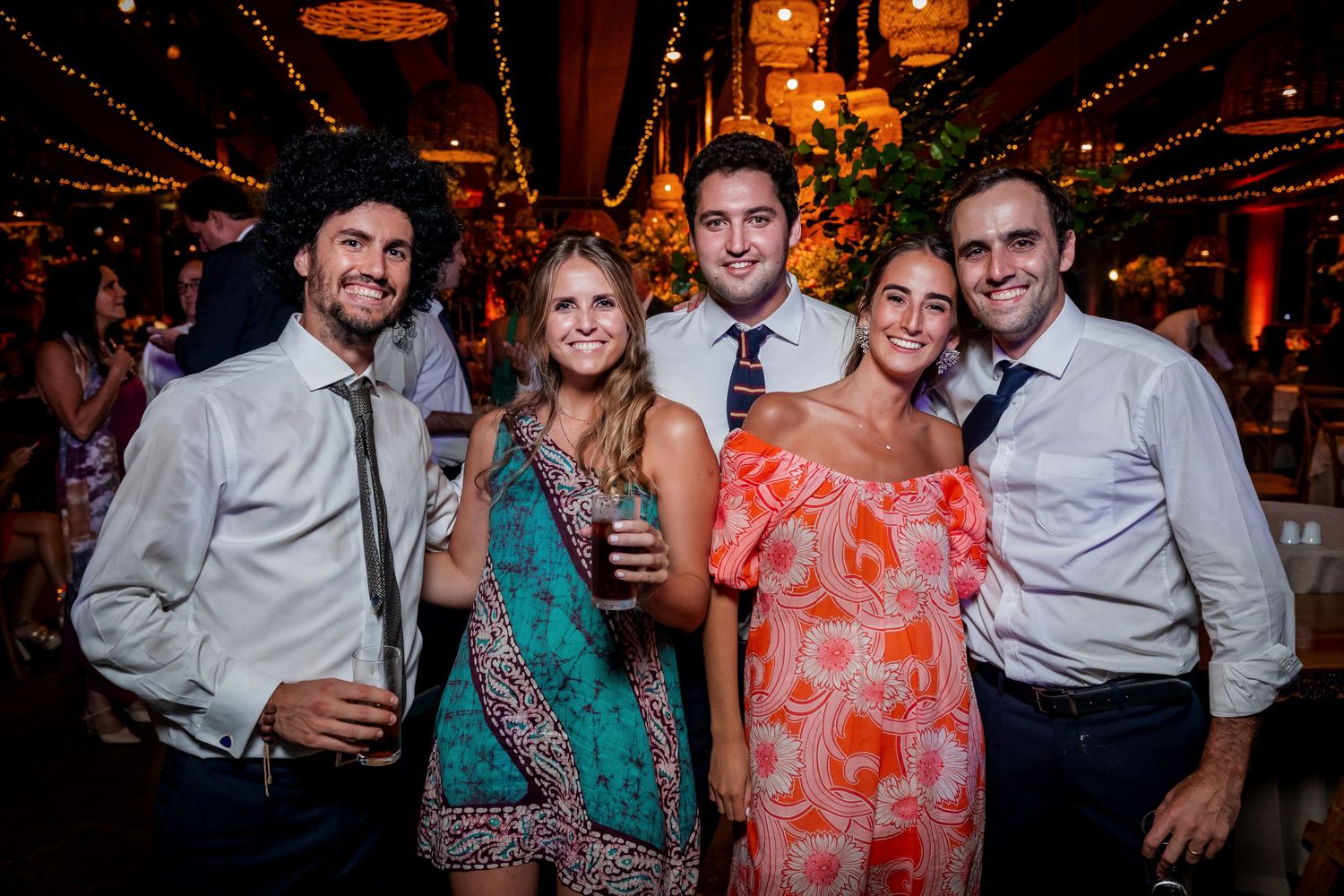 Five elegantly dressed wedding guests pose and smile together at a warmly lit wedding reception venue.
