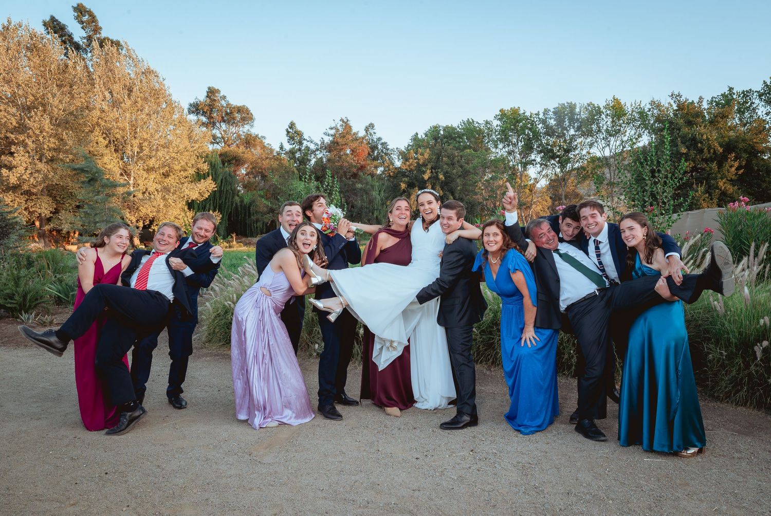 Wedding party celebrating outdoors as groomsmen lift the bride while guests pose playfully.