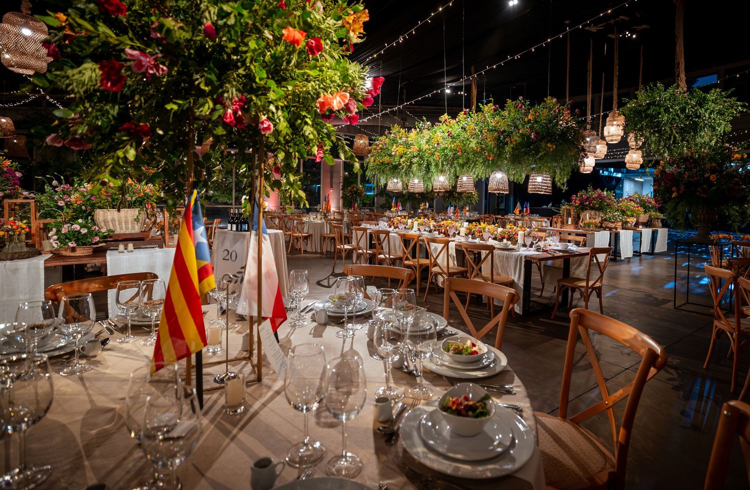 Elegant outdoor wedding reception with round tables, floral arrangements, string lights, and lush greenery at night.
