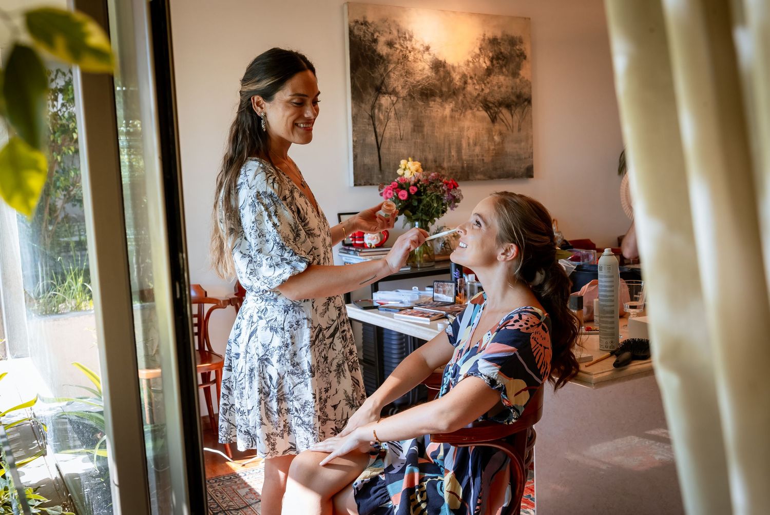 Two women in floral robes share a joyful moment with flowers during bridal preparations.