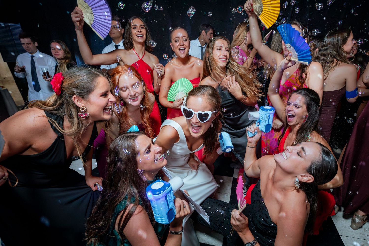 Bride in white outfit and heart sunglasses celebrating with guests on wedding dance floor.