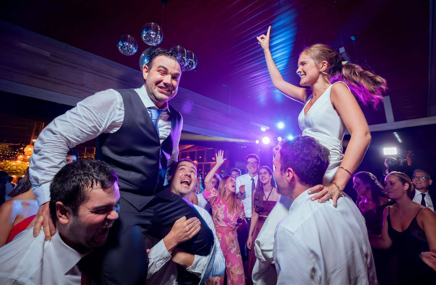 Bride and groom lifted on shoulders by wedding guests during a lively celebration on the dance floor.