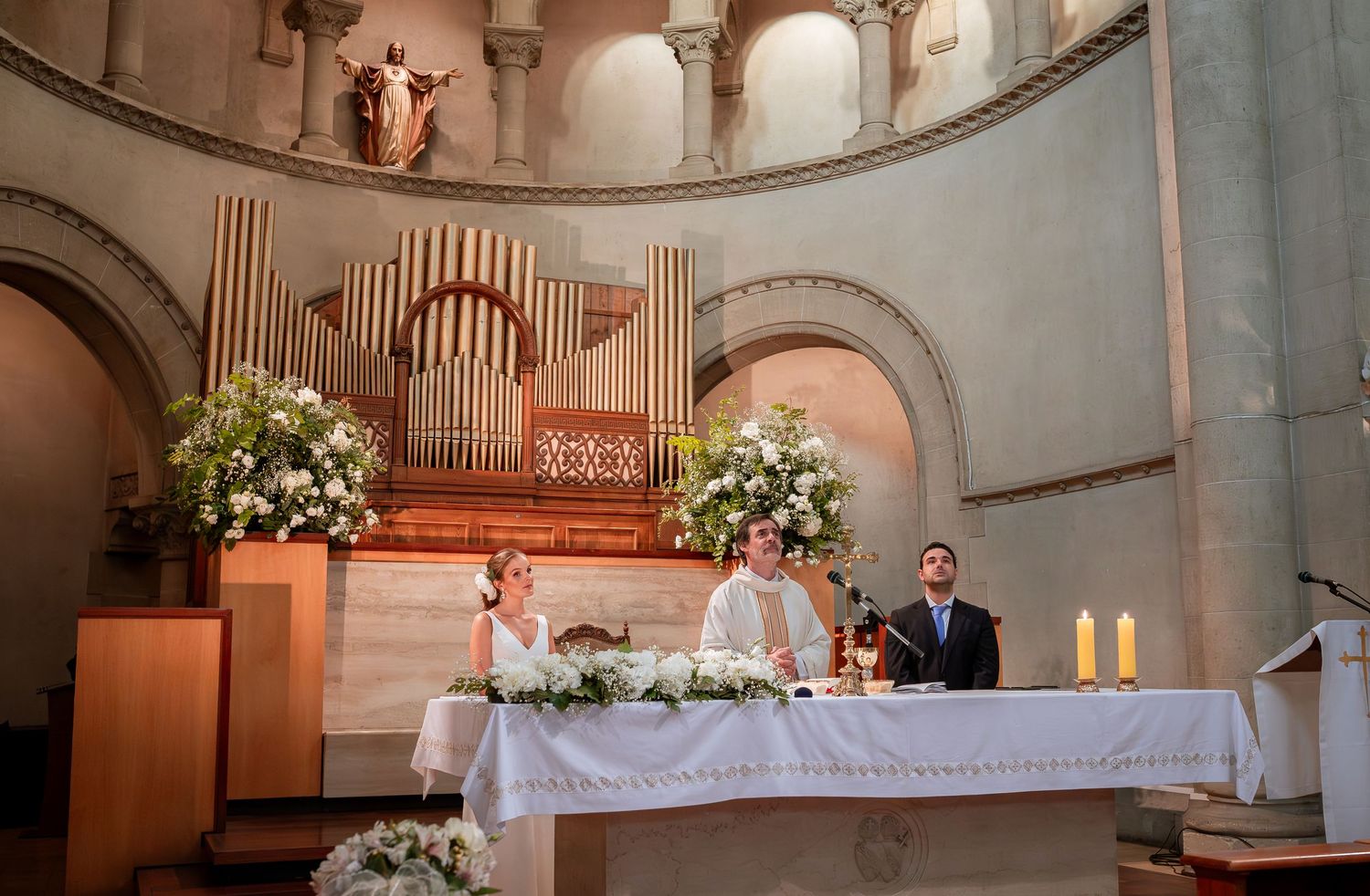 Bride and groom sit at altar with priest during Catholic wedding ceremony in elegant church with pipe organ.
