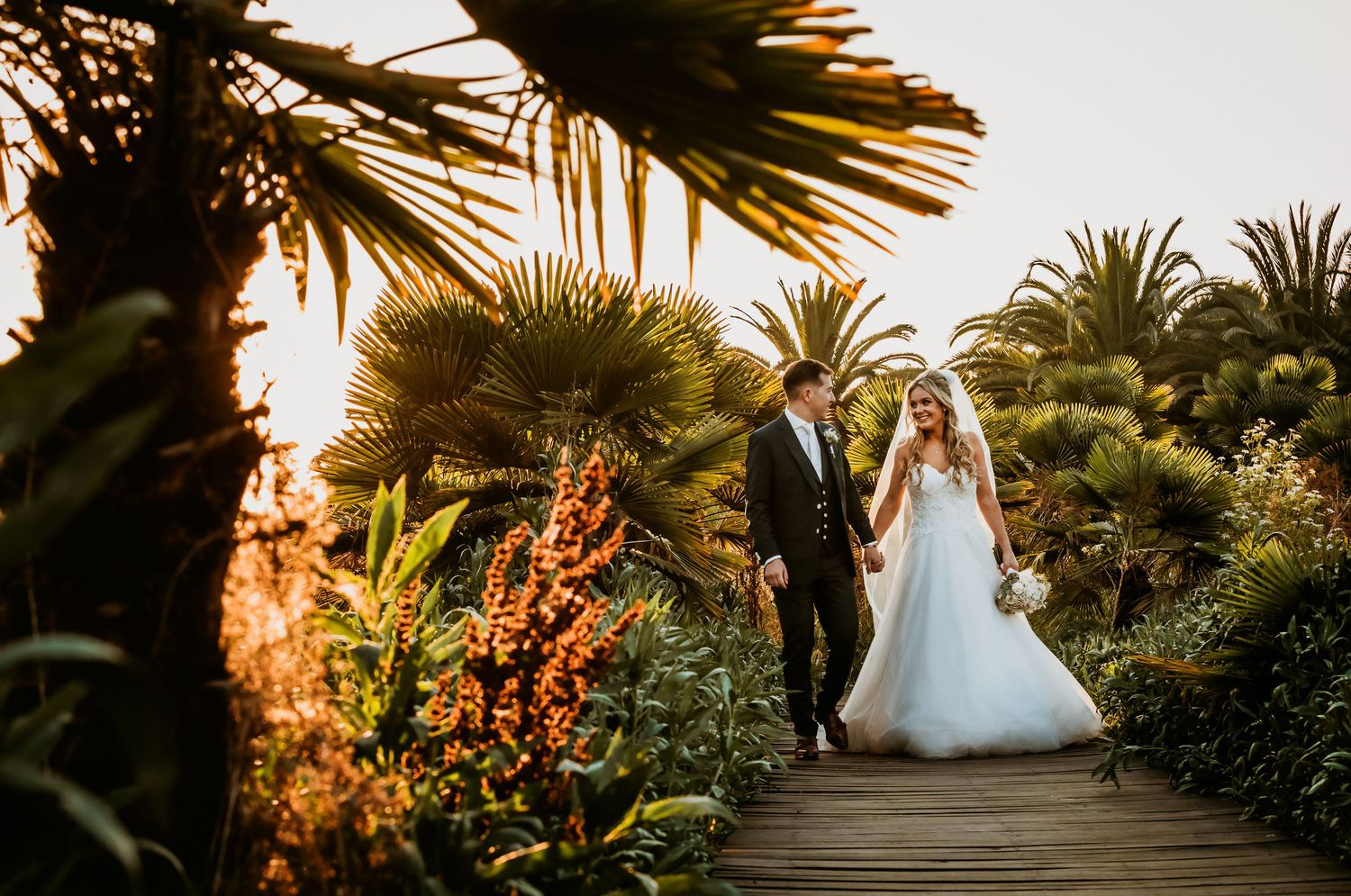 Bride and groom strolling along a wooden boardwalk through a vibrant tropical garden at golden hour.