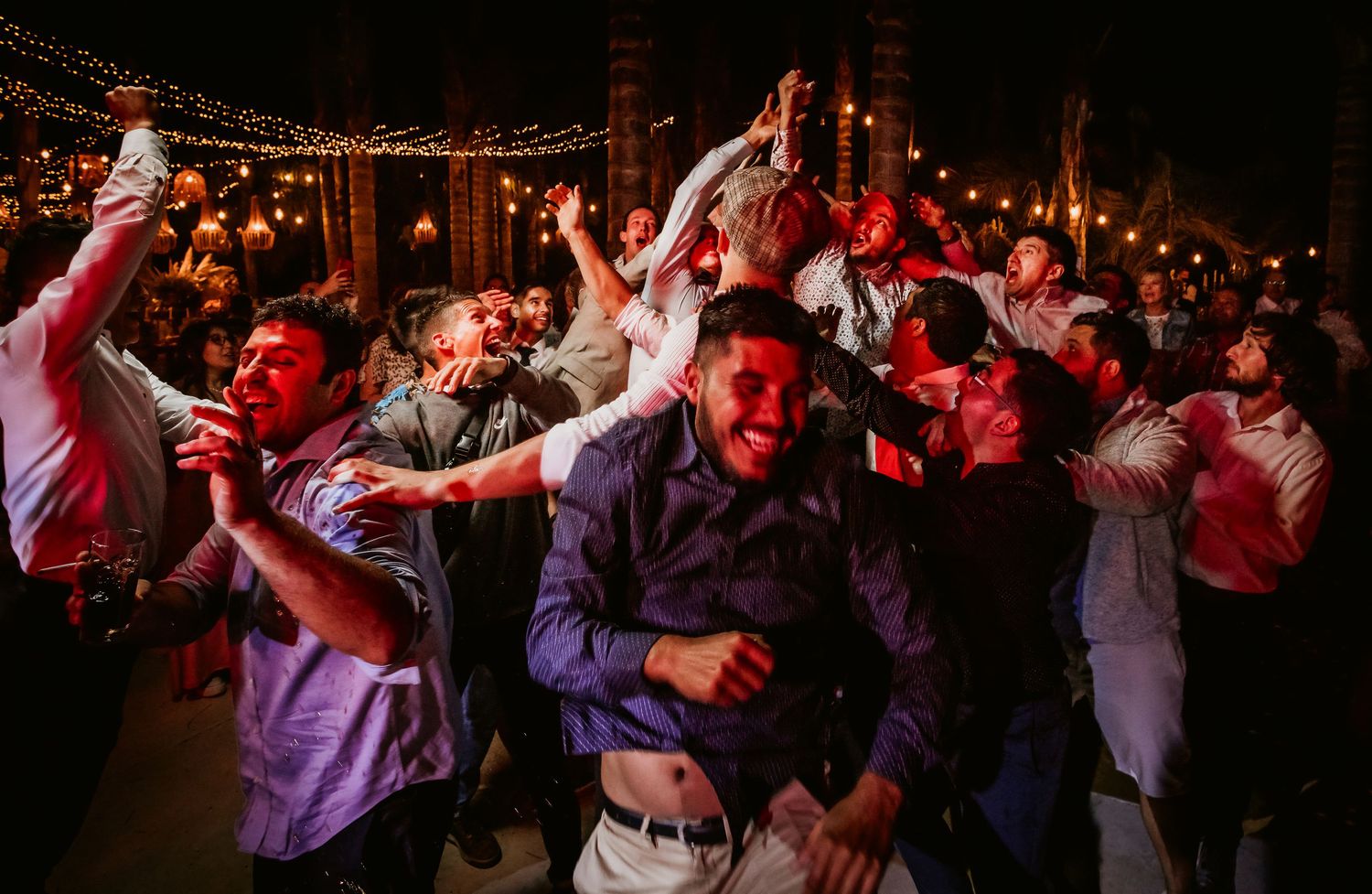 Energetic wedding reception dance floor with guests celebrating and dancing under warm string lights at night.