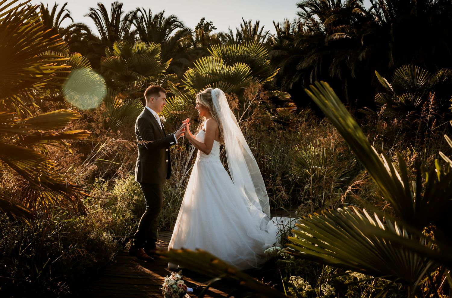 Bride and groom hold hands in sun-drenched tropical garden, bride in flowing white gown with veil.
