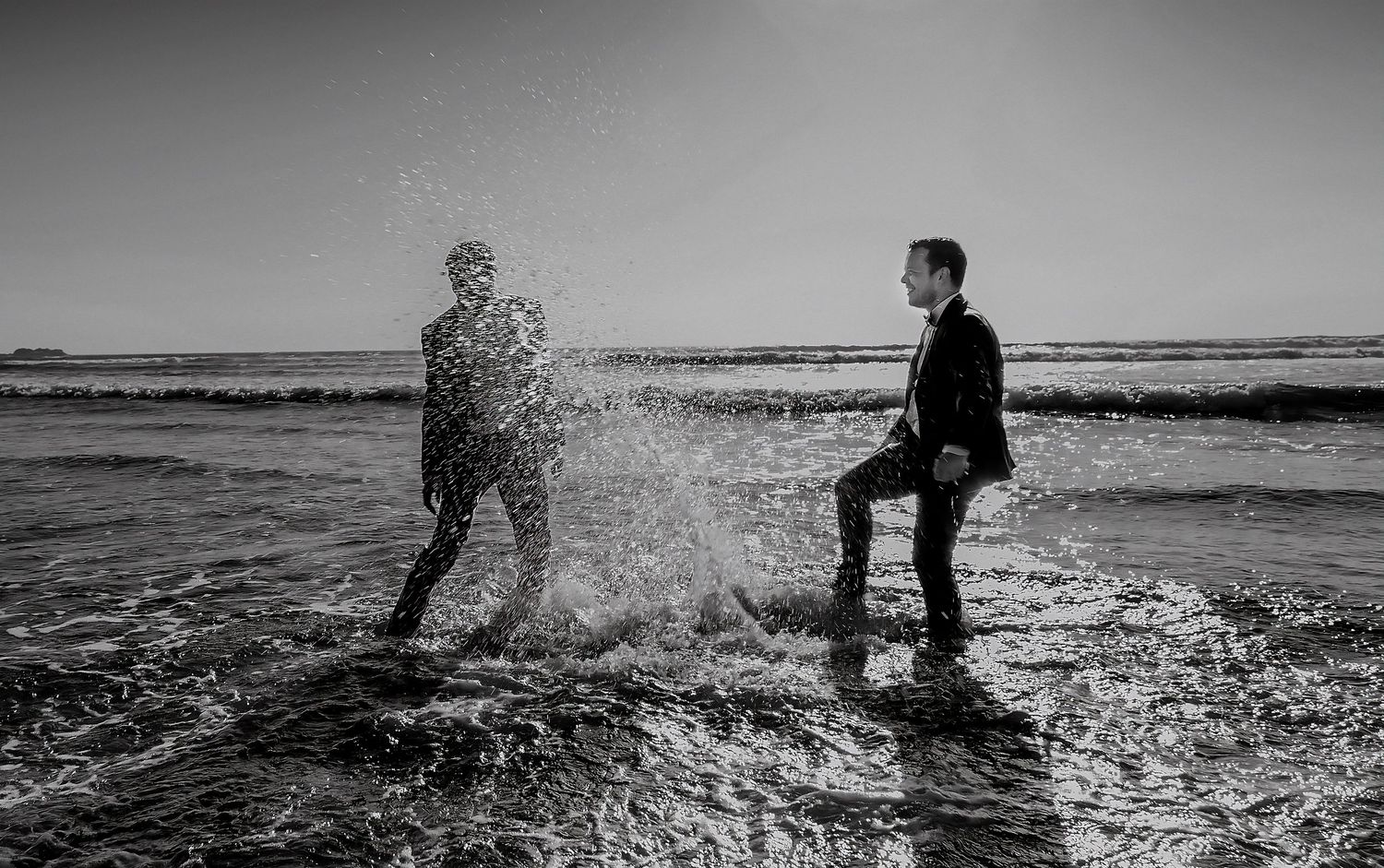 Black and white photo of two grooms, one kicking a dramatic water splash toward the other on a beach.
