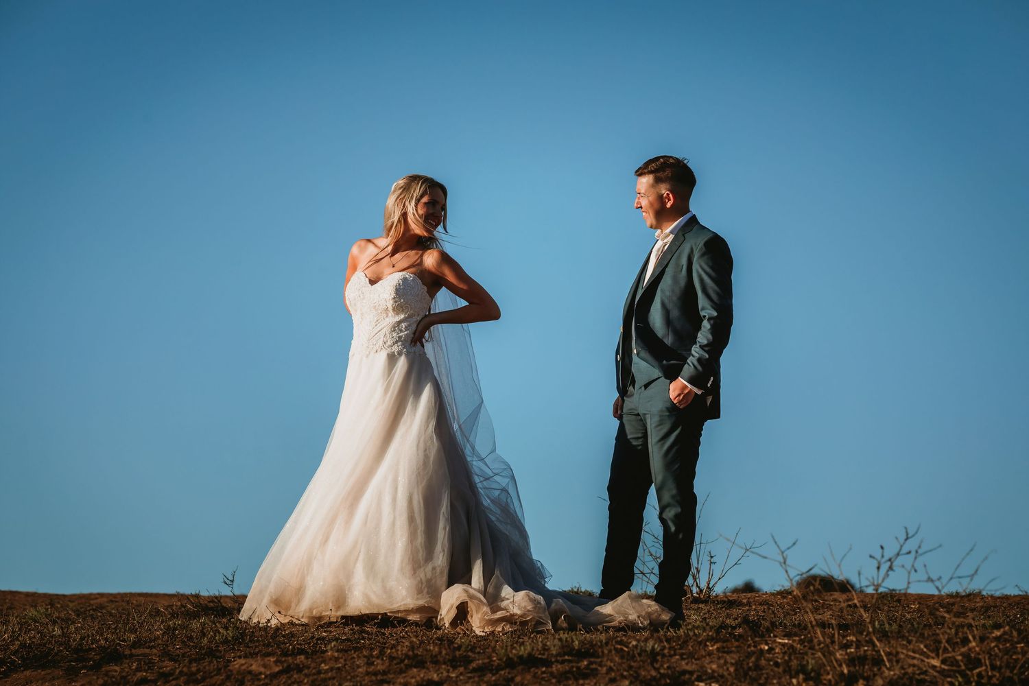 Bride and groom facing each other on a hilltop under a vivid blue sky.