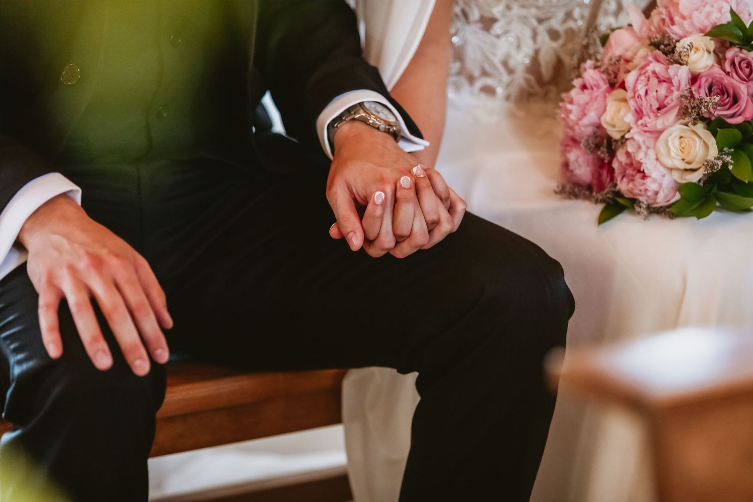 Bride and groom holding hands at wedding ceremony with pink bouquet nearby.