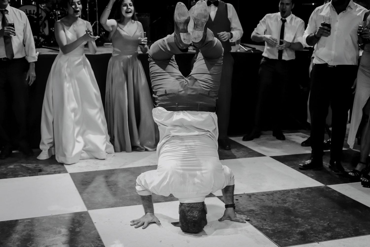 Guest performs energetic breakdance headstand on checkered floor as wedding party cheers around him.