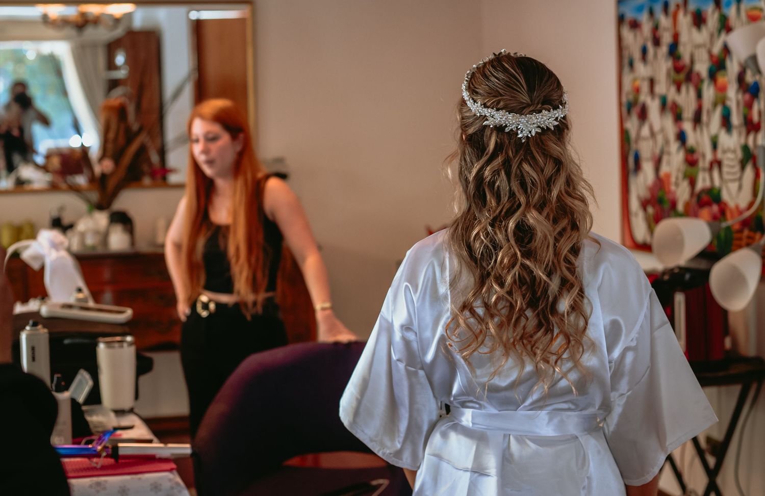 Bride in white robe with floral headpiece getting hair styled before wedding.