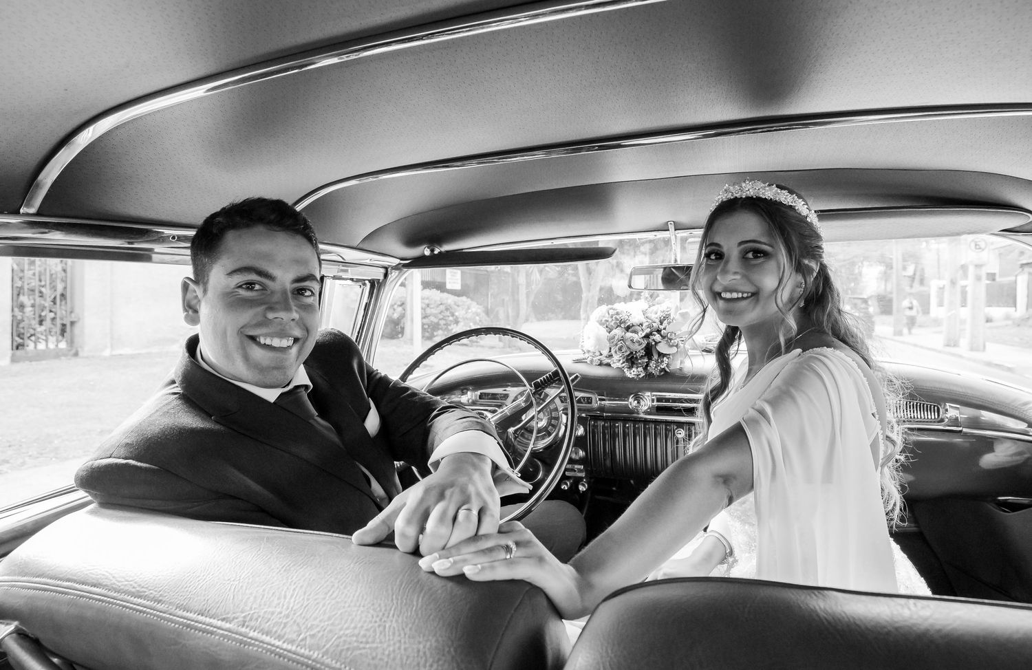Black and white photo of smiling bride and groom sitting inside a vintage classic car on wedding day.