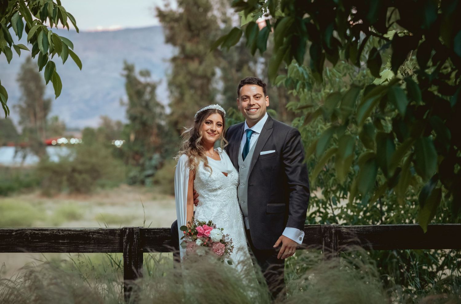 Smiling bride and groom pose together outdoors with mountains and greenery in the background.