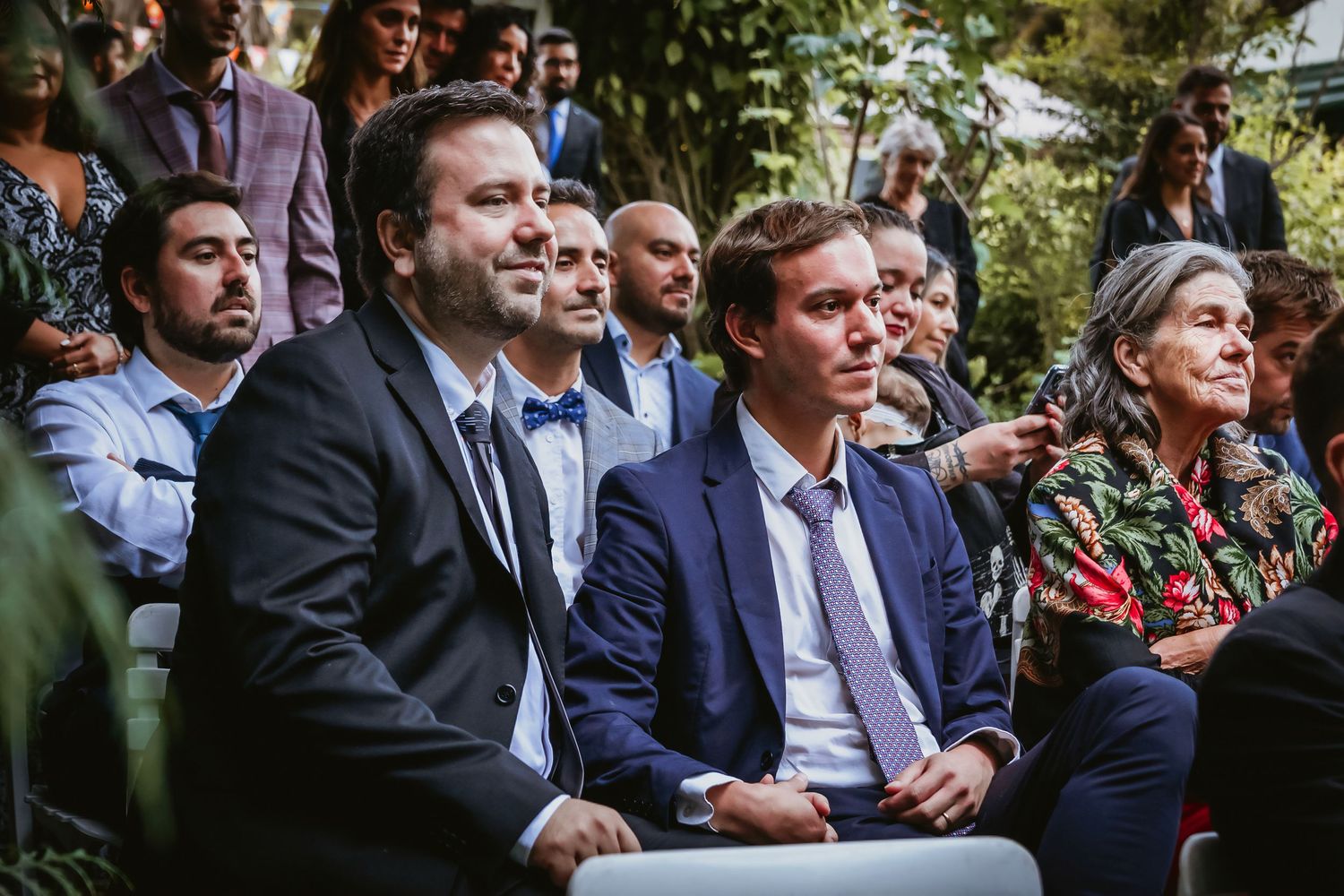 Elegantly dressed wedding guests seated in white chairs watching an outdoor garden ceremony with lush greenery.
