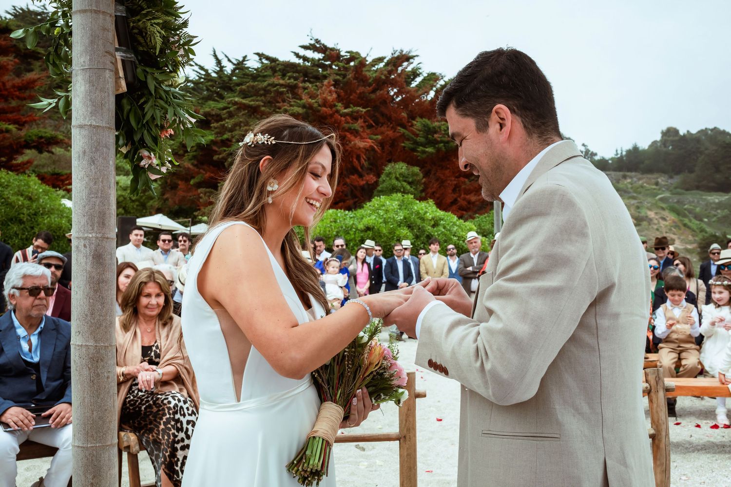 Bride smiles while placing wedding ring on grooms finger during sunny outdoor beach ceremony.