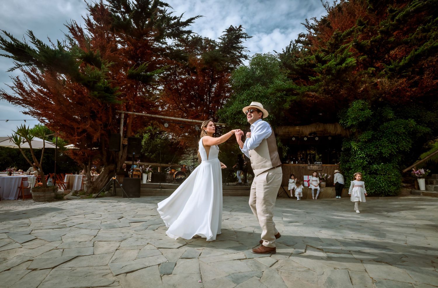 Bride in white gown and groom in hat twirl together during their first dance at an outdoor wedding reception.