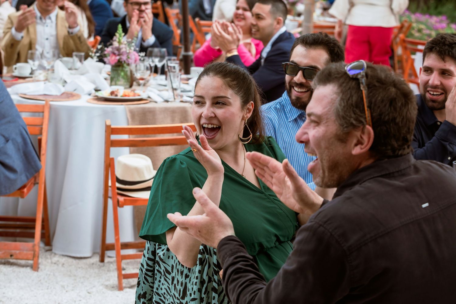 Woman in green dress laughs and claps with guests at a lively outdoor wedding reception dinner.