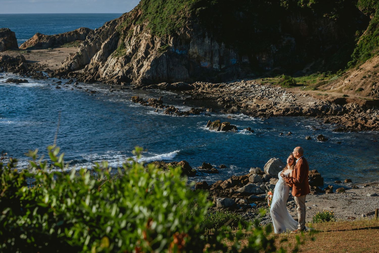 Couple in wedding attire embracing on a rocky coastal cliff overlooking turquoise ocean waves and rugged shoreline at sunset.