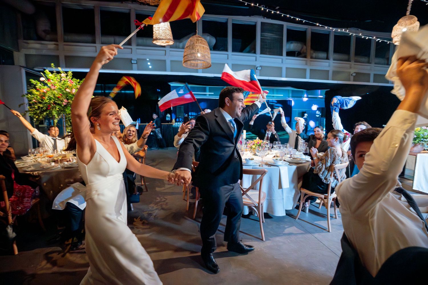 Newlyweds dancing at wedding reception entrance while guests wave colorful flags in decorated venue.