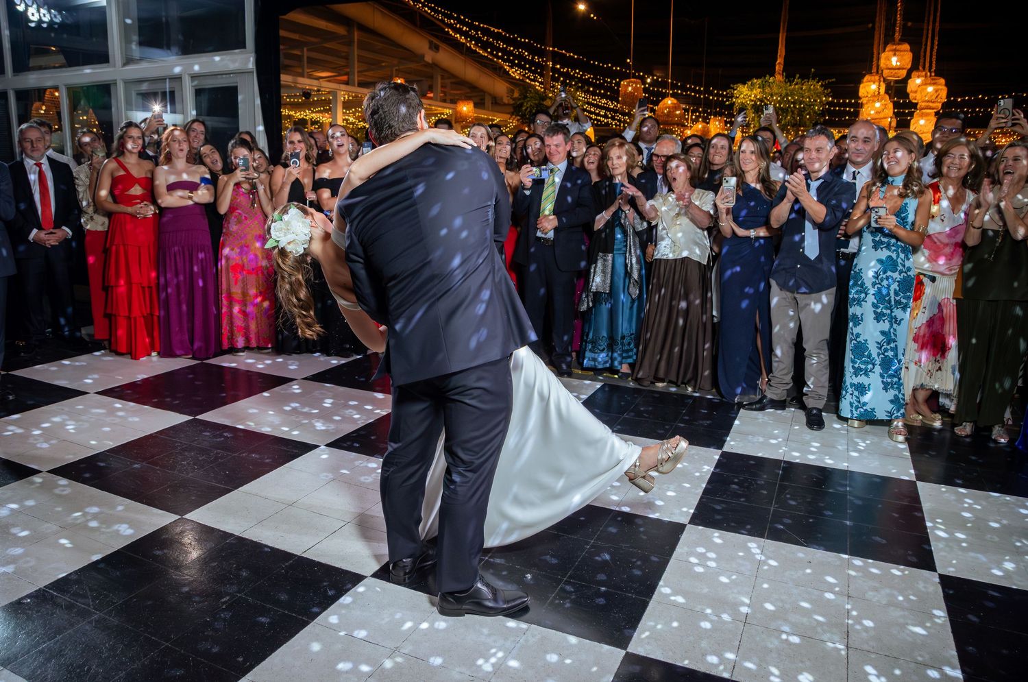 Groom lifts bride during their first dance on a checkered dance floor at an elegant outdoor wedding reception.
