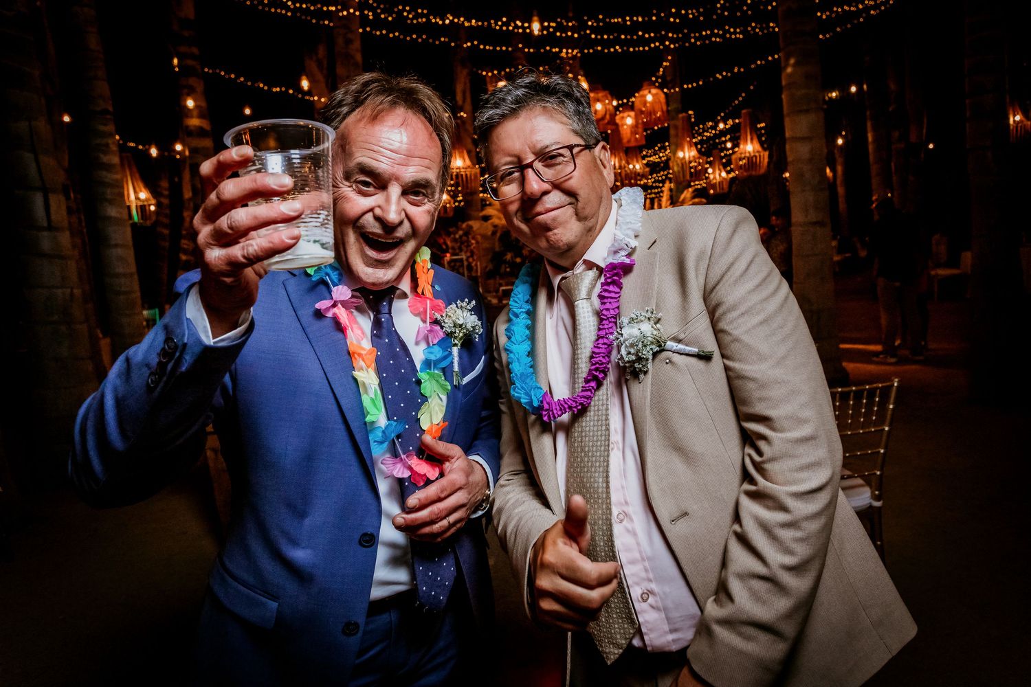 Two men in suits wearing flower leis raising glasses in a toast at outdoor wedding reception.