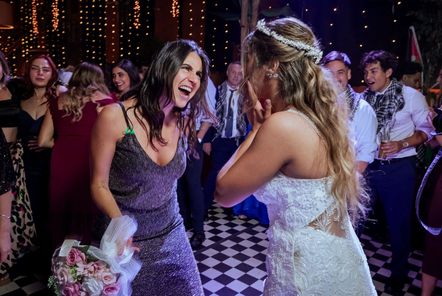 Bride in white gown laughs joyfully with guest in purple dress at wedding reception.